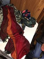 Three scarves laid out on a table including a long red-orange striped one, a botanical patterned one, and a dark multicolor one with pink and purple. Part of a black and white floral blouse also visible.