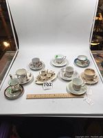 Photo showing seven demitasse teacups on matching saucers arranged around a miniature tea set on an oval tray.