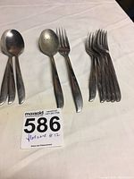 Photo showing a group of spoons and forks arranged on a white cloth. Includes 3 large spoons, 1 smaller spoon, and 8 forks, all matching in design.
