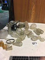 Assortment of clear glass textured and patterned lamp parts laid out on a table including globe shades and finials with some metal parts.