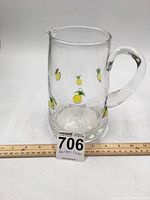 Front view of clear glass pitcher with painted yellow pineapples and handle. Ruler shows approximate height of 9 inches.