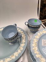 Photo showing several light blue Wedgwood teacups stacked on plates with white grapevine and floral relief decoration on rims.