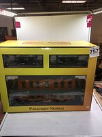 Front view of yellow box containing Rail King Passenger Station model, showing display windows revealing miniature building and fencing inside.