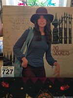 Front cover of Carly Simon's No Secrets vinyl album showing Carly Simon in blue attire with brown hat and black fence background.