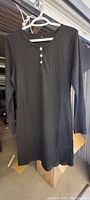 Single black ribbed knit dress hung on white plastic hanger showing front view with three white buttons at crew neckline