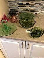 Overview of assorted colored glassware on countertop including large green serving plate with handle, multiple green bowls with patterns, and smaller pink and amber decorative glass pieces.