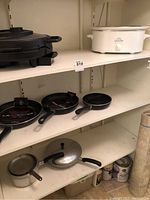 Shelf showing two black frying pans with red and white Logostina branding, Hamilton Beach indoor grill, and other cookware items beneath.
