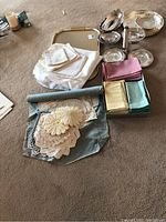 Overview photo showing assorted folded linens including white tablecloths, doilies, and several silver plated serving trays on carpet floor.
