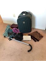 Green carry-on suitcase with Charlie Sport emblem, cane, multiple scarves, and clutches laid out on floor