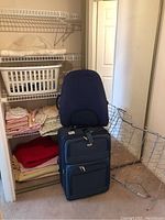 Photo showing folded linens including bed sheets and fleece blankets on shelves with white plastic laundry basket and navy blue soft suitcase in front