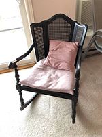 Front-right angle of wooden rocking chair showing rattan back and seat, curved top backrest, turned armrests and legs, and pink seat cushion