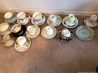 View of all 12 assorted tea cups and matching saucers arranged in two rows on carpeted floor