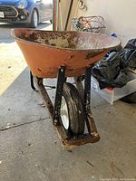 Front angled view of orange metal wheelbarrow with wooden handles and black metal frame, showing rust and wear on tray and some debris inside.