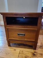 Front view of brown side table showing drawer with metal handle and open storage compartment with cable outlet hole.
