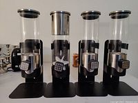 Front view of 4 candy dispensers in a row showing clear cylindrical containers and black push-button dispensing mechanisms on black bases.