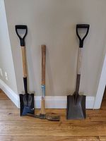 Front view of pickaxe center with two square head shovels on either side against wall and floor background.