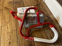 Folded aluminum emergency fire escape ladder with red and white frame, resting on wooden floor showing attached red straps and packed instruction manual.