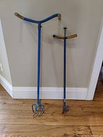 Full view of two blue gardening tools with wooden handles against a wall on hardwood floor.