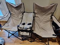 Two beige folding lawn chairs side by side with black trim, each having an armrest with built-in drink holders. Chairs have metal framing and fabric seats with visible usage signs.