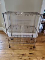 Front view of a three-tier metal wire shelving unit with caster wheels, placed on a wooden floor.
