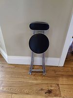 Mini stool folded, showing round seat and small backrest with black upholstery and white piping