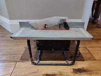 Full view of the portable table saw showing metal table surface, blade with protective guard, and tubular metal legs. Some dust and wear visible.