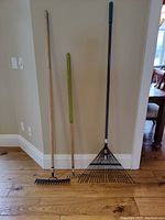 Three garden rakes standing against a wall on a wooden floor: large fan rake with blue handle, medium straight back rake, mini straight back rake with green handle.