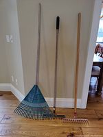 Full view of the three garden rakes standing upright against a wall on wooden floor showing size and color differences.