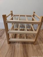 Small wooden wine bottle rack displayed on a floor against a wall, showing open construction and horizontal bars for holding bottles.