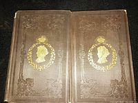 Both antique volumes closed, showing brown cloth covers with embossed ornate floral border and gold profile silhouette emblem with laurel wreath and sunburst.