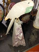 Front and side view of carved quartz parrot showing the green beak, white quartz body, and metal claws perched on an amethyst geode base.