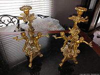 Pair of gold-tone dore bronze candle holders each featuring multiple cupids holding birds and floral motifs, placed on black reflective surface.