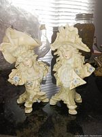 Photo showing two German Dresden porcelain figures, a man and a woman, both wearing elaborate period costumes with high detail and blue floral relief decoration.