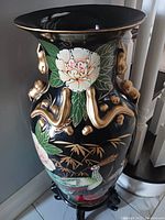 Photo showing black oriental vase with large white flower, gold raised handles, and a bird design, standing on black carved wooden stand.