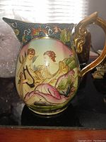 Side view of decorative hand painted ceramic pitcher with classical woman and cherub scene, detailed gold handle, and ornate Moriage texture.