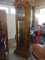 Full view of oak grandfather clock standing 83 inches tall with carved crown and glass panel door in a room with other furniture and tools.