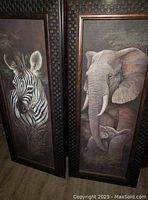 Photo showing both framed animal prints side by side; zebra on the left, elephant and calf on the right. Frames are dark wood with woven or checkerboard style detailing around edges.