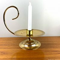 Close-up of brass chamber style candle holder with white taper candle on wooden surface, showing curved handle, round base, and circular drip plate.