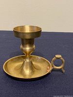 Full view of the brass chamberstick candle holder on a navy cloth background, showing the bowl-shaped candle holder top, slender neck, wide base plate, and round handle.