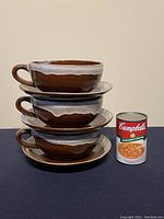Stacked view of three large ceramic soup mug bowls with matching saucer plates, next to a Campbell's soup can for size reference