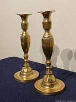 Pair of tall, solid brass candlesticks shown from a front angle. Both have a polished golden finish and classic shape with broad, rounded bases and flared tops for holding candles.