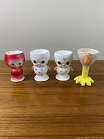 Four vintage egg cups arranged side by side on a wooden surface, showing front view of chick and chicken designs.