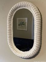 Front angled view of the oval vintage white wicker wall mirror showing the woven wicker texture and reflective glass surface in a room setting.