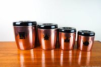 Four rosegold metal canisters with black lids and plastic knobs, labeled for Flour, Sugar, Coffee, and Tea respectively, arranged in descending size order on a wood surface.