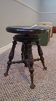 Photo of the antique wood adjustable piano stool showing the round seat, turned legs, and claw feet on carpeted floor with vintage suitcases in background.