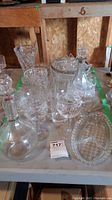 Photo showing multiple glass items including decanters, wine glasses, and serving dishes arranged on a table in a garage setting.