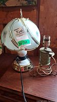 Photo showing two lamps on wooden surface. One lamp has cream-colored glass shade with floral decoration and brass base. Second lamp has metal painted decorative body, crank handle, no shade.