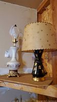 Two Mid-Century Modern lamps on wooden shelf, one with beige fiberglass shade decorated with stars, black base with gold star embellishments, other white ceramic lamp without shade