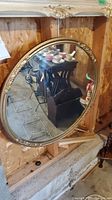 Round gold-tone framed wall mirror leaning against a wooden wall, showing reflection of surrounding antiques and furniture.