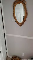 Gold framed oval mirror with ornate carved detailing hung on a wall above a wooden floating shelf.